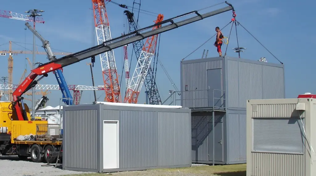 Jobs bei MBN Modulwelt Arbeiter steht auf einem Bürocontainer, der mit Hilfe eines Kranwagens auf einem anderen Containermodul plaziert wird