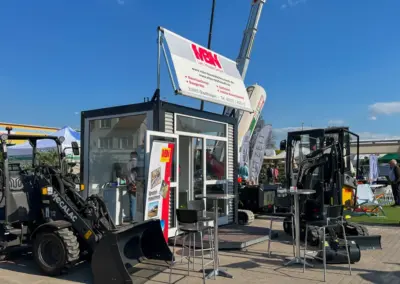 Messestand mit Containerbüro und ausgestellten Baumaschinen (Bagger und Radlader) auf einer Outdoor-Fachmesse unter blauem Himmel.