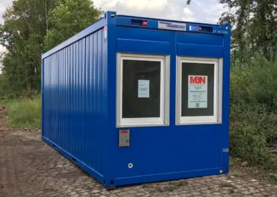 Blauer Baucontainer von MBN-Modulwelt mit zwei Fenstern, aufgestellt auf einem gepflasterten Weg in grüner Umgebung.