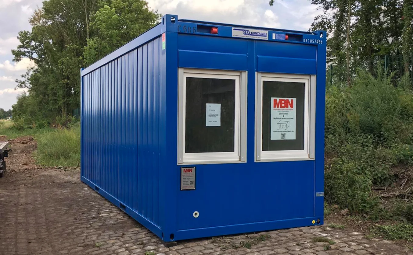 Blauer Baucontainer von MBN-Modulwelt mit zwei Fenstern, aufgestellt auf einem gepflasterten Weg in grüner Umgebung.
