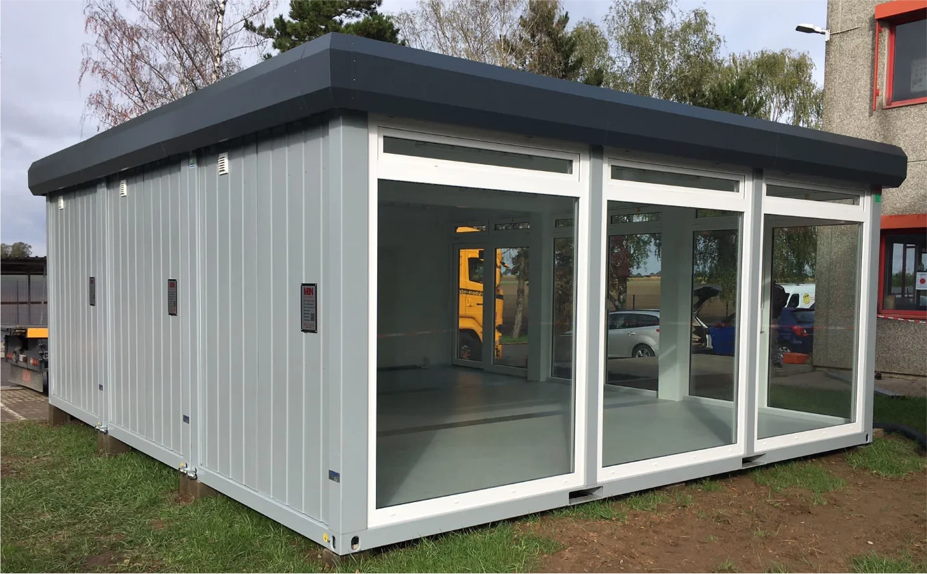 Moderner MBN Modulwelt Bürocontainer mit großen Glasfronten und heller Innenfläche, aufgestellt im Außenbereich neben einem Gebäude.