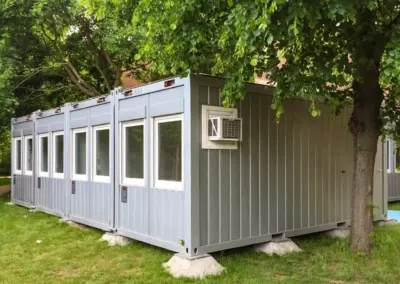 Reihe von modularen Containern mit Fenstern und Klimagerät, aufgestellt im Grünen unter Bäumen, geeignet für Gastronomie oder Nutzung im Außenbereich.