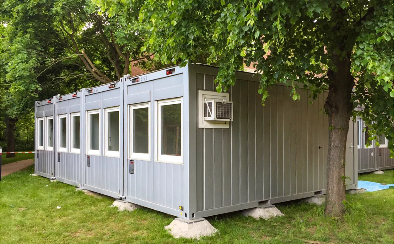 MBN Modulwelt Bürocontaineranlage silber mit Klimaanlage Reihe von modularen Containern mit Fenstern und Klimagerät, aufgestellt im Grünen unter Bäumen, geeignet für Gastronomie oder Nutzung im Außenbereich.