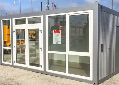 Moderner Verkaufscontainer mit großer Glasfront und Eingangstür, aufgestellt auf einem Betriebsgelände.