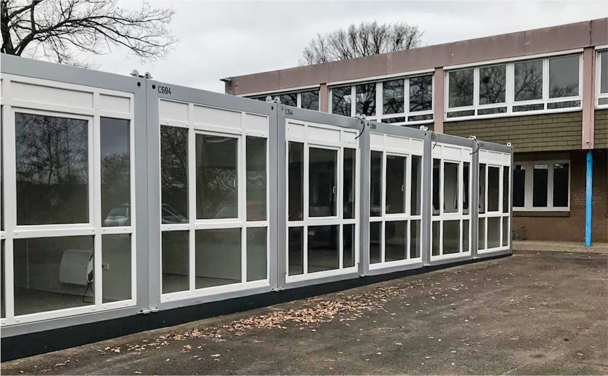Reihe von Schulcontainern mit großen Fensterflächen, aufgestellt vor einem Schulgebäude als temporäre Klassenräume.