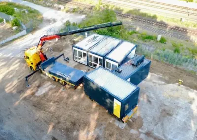Eine Luftaufnahme aus der Vogelperspektive zeigt eine Baustelle in der Nähe von Bahngleisen. Ein gelber Kran-LKW (mit MBN-Branding) setzt gerade den Dachbereich einer modularen Containeranlage zusammen, die aus mehreren blauen Modulen mit weißen Dächern und Glasfronten besteht. Ein weiterer dunkelblauer Lagercontainer steht im Vordergrund auf dem Schotterplatz. Im Hintergrund fahren Züge auf den Gleisen.