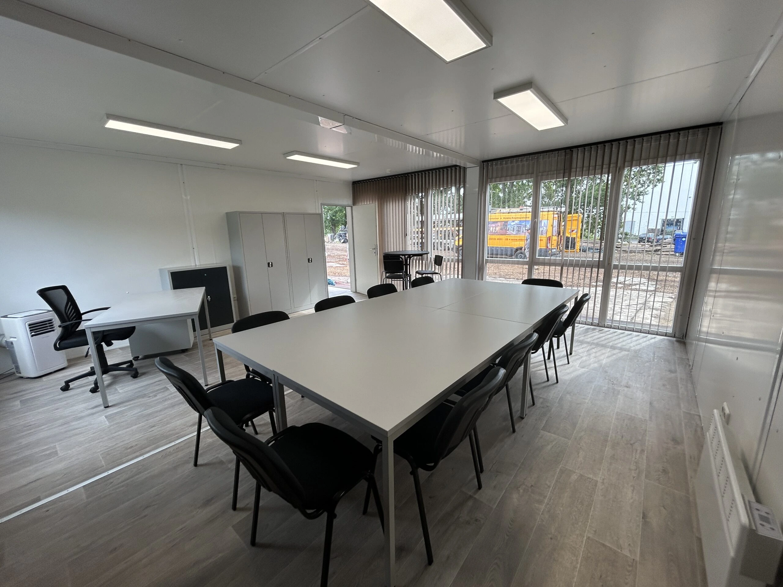 Heller, weitläufiger Konferenzraum in einer Containeranlage mit großem weißem Meetingtisch, schwarzen Konferenzstühlen, Boden in Holzoptik, bodentiefen Glasfronten mit Vertikal-Lamellen sowie einem zusätzlichen Arbeitsplatz und Büroschränken.