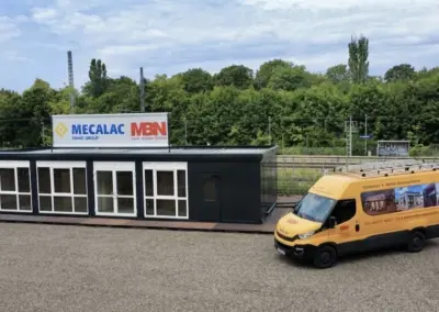 Luftaufnahme einer modernen, einstöckigen, schwarzen Containeranlage mit großzügigen Glasfronten und Glastüren, die auf einem Schottergelände steht. Oben auf der Anlage ist ein großes Werbebanner mit den Logos von "MECALAC FAYAT GROUP" und "MBN" befestigt. Rechts daneben steht ein gelber Iveco Daily-Lieferwagen mit MBN-Branding ("MBN - Container + Mobile Raumsysteme"), einer Website und Telefonnummer sowie kleinen Bildern von Containern auf der Seite. Der Hintergrund zeigt grüne Bäume und Bahngleise unter einem bewölkten Himmel.