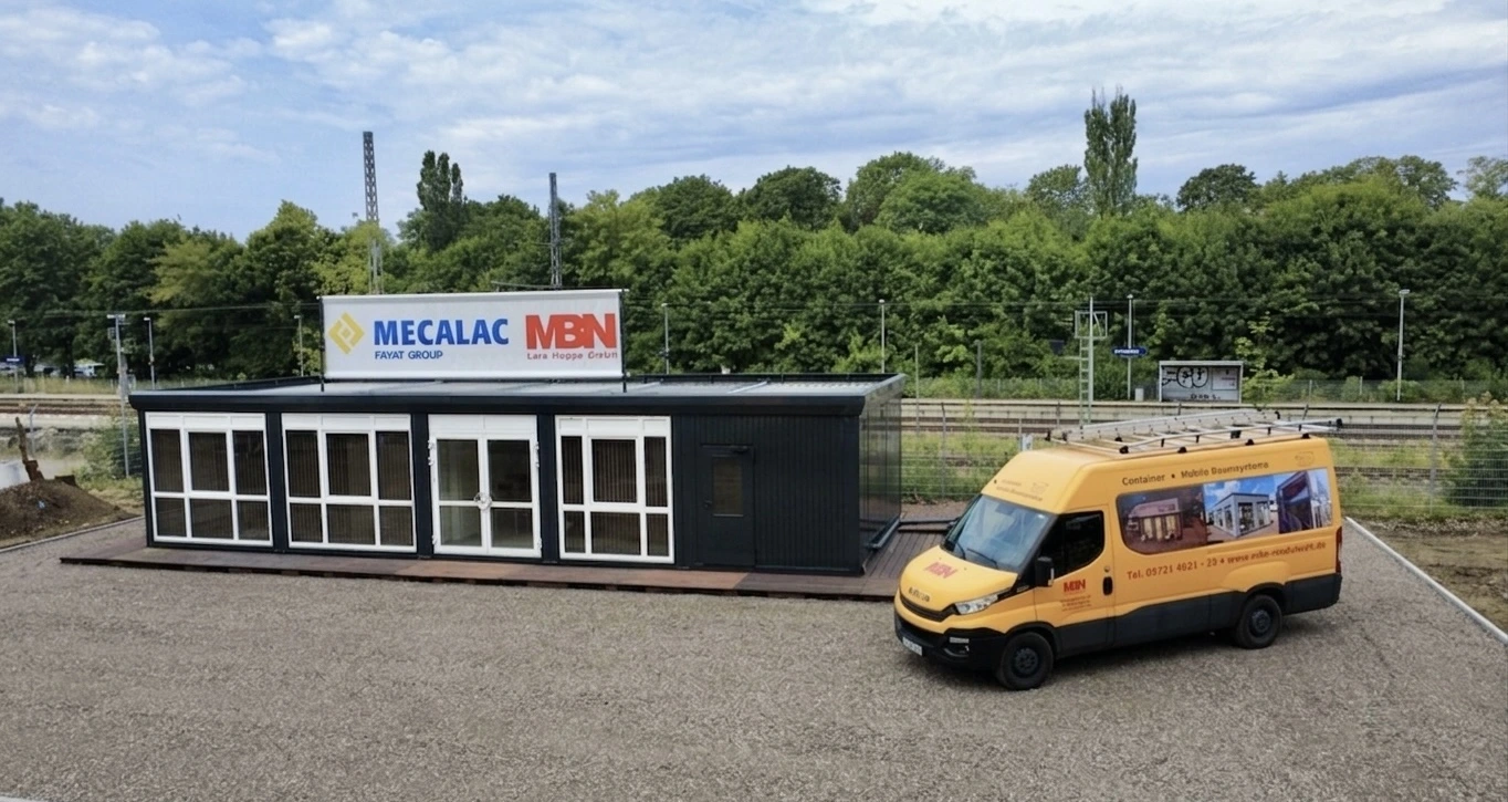 Luftaufnahme einer modernen, einstöckigen, schwarzen Containeranlage mit großzügigen Glasfronten und Glastüren, die auf einem Schottergelände steht. Oben auf der Anlage ist ein großes Werbebanner mit den Logos von "MECALAC FAYAT GROUP" und "MBN" befestigt. Rechts daneben steht ein gelber Iveco Daily-Lieferwagen mit MBN-Branding ("MBN - Container + Mobile Raumsysteme"), einer Website und Telefonnummer sowie kleinen Bildern von Containern auf der Seite. Der Hintergrund zeigt grüne Bäume und Bahngleise unter einem bewölkten Himmel.