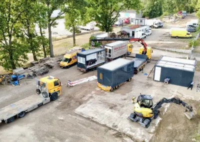 Eine Luftaufnahme zeigt ein Betriebsgelände, auf dem Arbeiter mobile Container-Raumsysteme montieren. Ein großer LKW-Kran hebt gerade ein Modul an, während ein Mecalac-Radlader Erdarbeiten durchführt. Mehrere dunkelgraue und ein gelber Service-Container stehen bereit, ebenso ein LKW-Sattelauflieger. Der Hintergrund zeigt gestapeltes Bauholz und einen Transporter.
