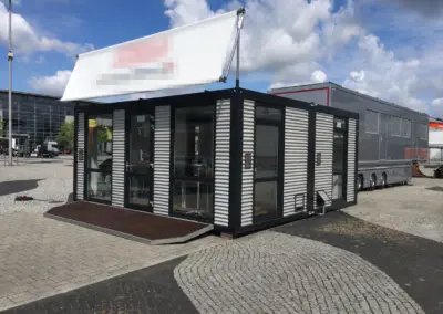 Moderner Bürocontainer mit schwarzer Rahmenkonstruktion, Glasfront und überdachtem Eingangsbereich auf einem Messegelände