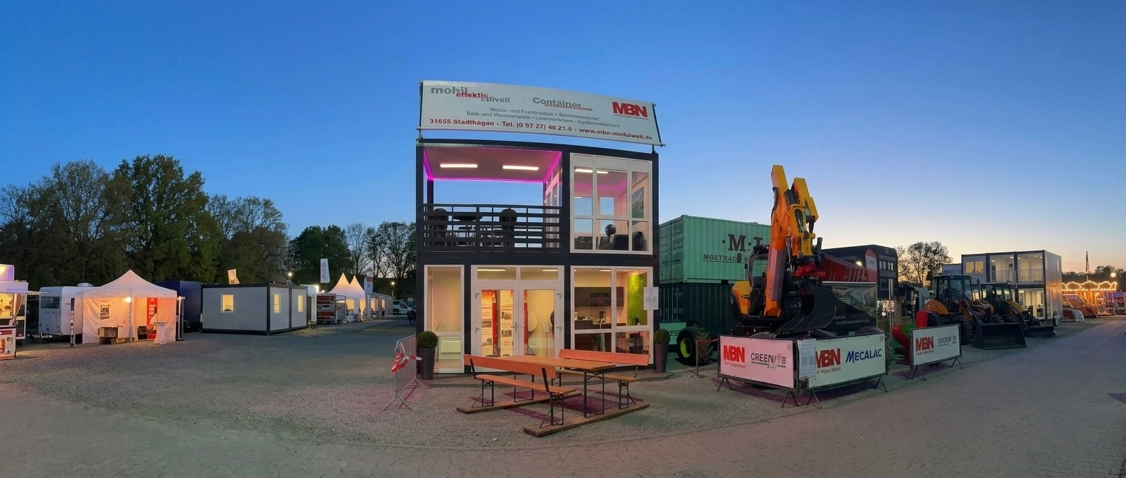 Moderne, zweigeschossige Messe-Containeranlage von MBN mit verglaster Front, stimmungsvoller LED-Beleuchtung, Dachterrasse und Werbebanner auf einem Freigelände bei Dämmerung.