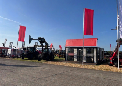 Mobiler Messe-Container mit großer Glasfront und rotem Vordach auf einem Ausstellungsgelände. Links stehen mehrere schwarze Traktoren und weitere Landmaschinen unter klarem blauen Himmel. Rote Fahnen sind gehisst.