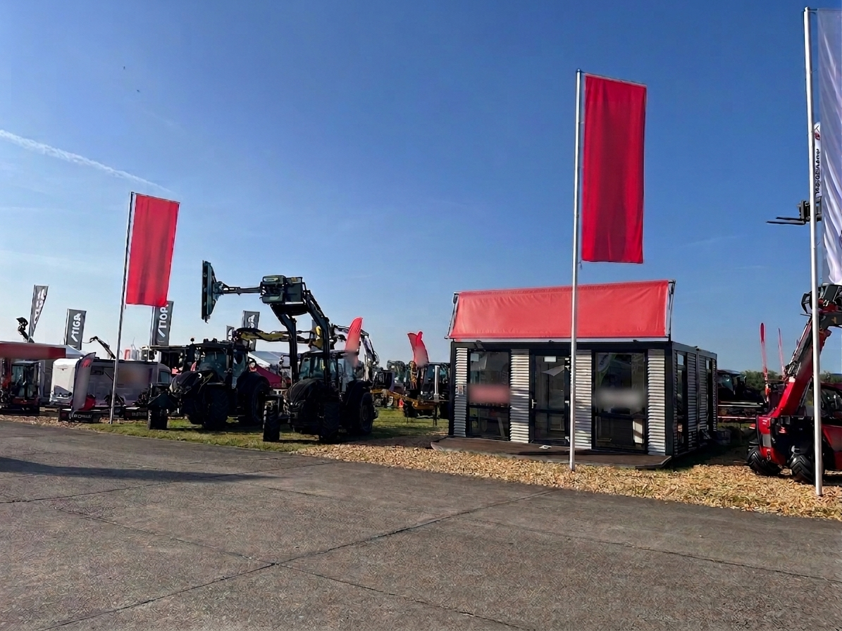 Mobiler Messe-Container mit großer Glasfront und rotem Vordach auf einem Ausstellungsgelände. Links stehen mehrere schwarze Traktoren und weitere Landmaschinen unter klarem blauen Himmel. Rote Fahnen sind gehisst.