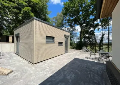 Freistehendes Tiny House von MBN mit hellgrau-beiger Holzoptik-Fassade, Flachdach, anthrazitfarbenen Fenstern und Überdachung auf einer gepflasterten Terrasse im Sonnenschein.