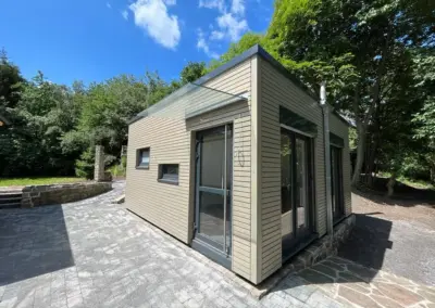 Modernes MBN Tiny House in Holzoptik mit großzügigen, bodentiefen Fenstern, Glaseingangsdach und Edelstahl-Schornstein auf einer gepflasterten Terrasse am Waldrand.