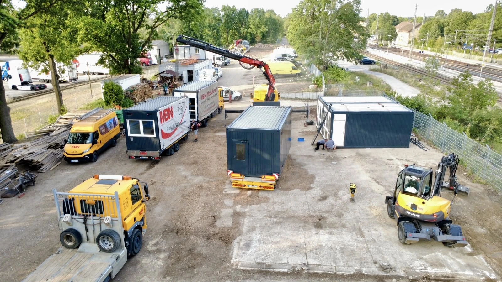MBN Modulwelt – Container Montage & Anlieferung Luftaufnahme der Montage von MBN Modulwelt-Containern mit LKW-Ladekran und Mobilbagger auf einem Baustellengelände.