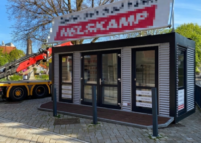 Moderner, modularer Showroom-Container mit großer Glasfront und Wellblechfassade auf einem gepflasterten Veranstaltungsgelände. Ein großes Werbebanner oben mit den Firmennamen. Links ist ein roter Autokran sichtbar, im Hintergrund Bäume unter blauem Himmel.