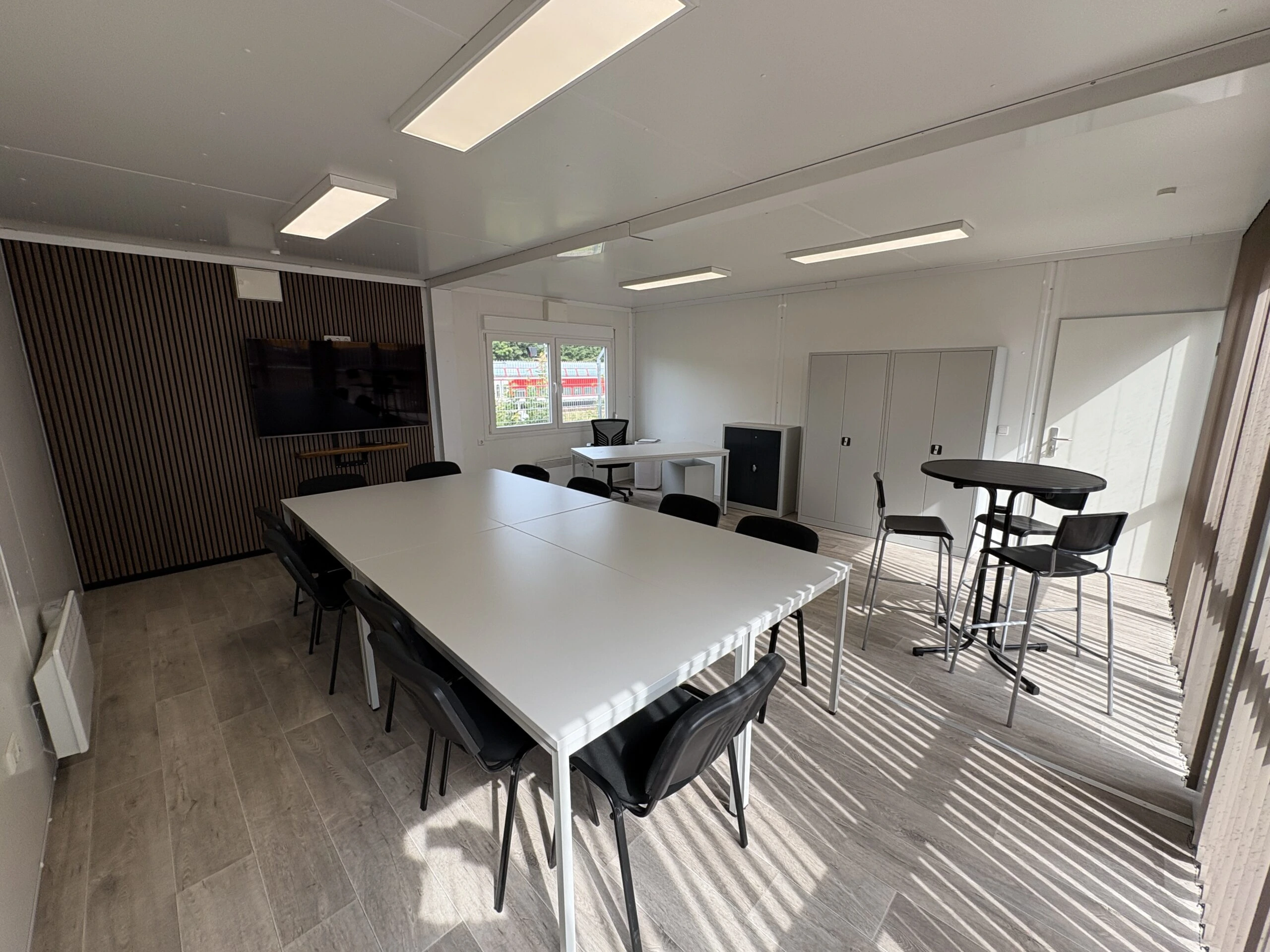 Moderner Konferenzraum in Containeranlage mit Meetingtisch, TV-Wand und Stehtisch-Lounge Perspektivische Innenansicht eines hellen Besprechungsraums in einer Containeranlage mit zentralem weißem Konferenztisch, schwarzer Bestuhlung, einer Akustikwand mit TV-Flachbildschirm, einem Stehtischbereich und Holzoptik-Boden mit sonnigem Lichteinfall.