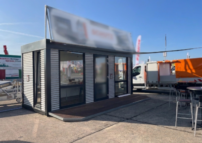 Mobiler Showroom-Container in Wellblech-Optik mit großer Glasfront und Holzterrasse auf einem Messe-Außengelände. Rechts stehen Barhocker und Tische, im Hintergrund sind ein weißer Lieferwagen und ein oranger Anhänger sichtbar. Das Werbebanner oben kann mit dem Firmeneigenen Logo versehen werden.