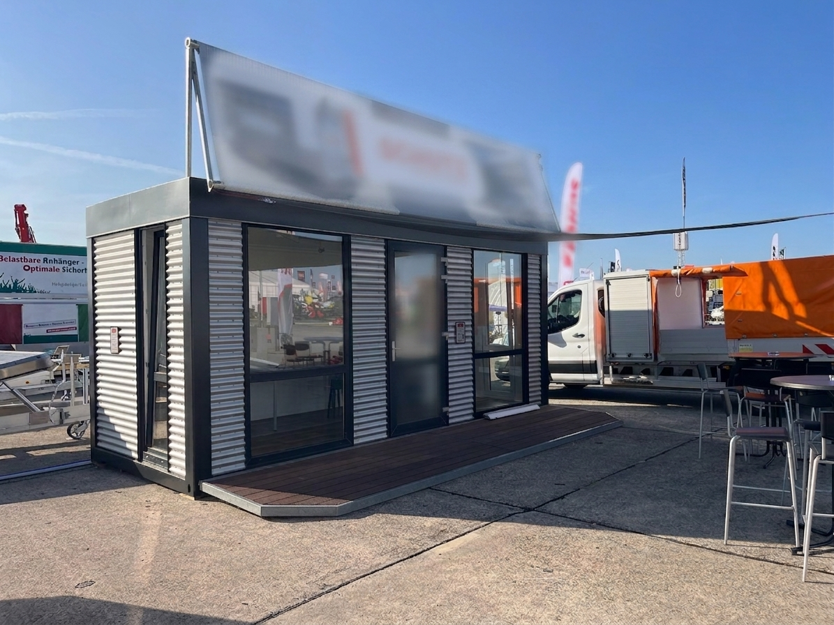 Mobiler Showroom-Container in Wellblech-Optik mit großer Glasfront und Holzterrasse auf einem Messe-Außengelände. Rechts stehen Barhocker und Tische, im Hintergrund sind ein weißer Lieferwagen und ein oranger Anhänger sichtbar. Das Werbebanner oben kann mit dem Firmeneigenen Logo versehen werden.