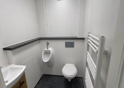 Detaillierte Innenansicht eines modernen, sauberen Toilettenraums in einer Containeranlage. Zu sehen sind ein Wand-WC mit Spülplatte, ein Urinal, ein Waschbecken mit Holzunterschrank, ein weißer Handtuchwärmer-Heizkörper, dunkler gesprenkelter Boden und Deckenbeleuchtung. Ideal für mobile Badezimmer und Sanitärcontainer-Lösungen.