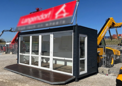 Anthrazitfarbener Event- und Showroom-Container von MBN mit weiß eingefasster Glasfront, dunklem Holzdeck und rotem Werbebanner auf einem Außengelände mit Baumaschinen.