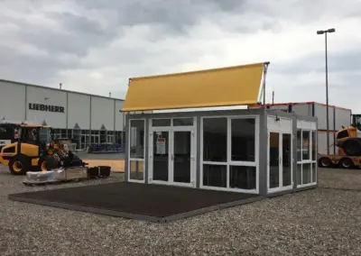silberner modularer Event- und Showroom-Container mit weiß gerahmten Panoramaglasfenstern, großer vorgelagerter Terrasse aus dunklem Holz und einem blanken gelben Werbebanner auf dem Dach.
