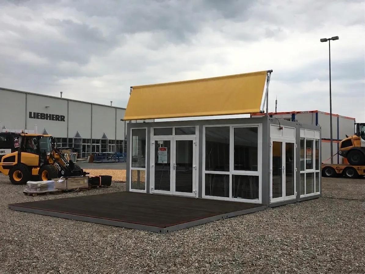 silberner modularer Event- und Showroom-Container mit weiß gerahmten Panoramaglasfenstern, großer vorgelagerter Terrasse aus dunklem Holz und einem blanken gelben Werbebanner auf dem Dach.