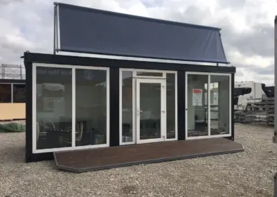 Muster-Eventcontainer von MBN mit schwarzem Rahmen, weiß gefassten Panoramaglasfenstern, Holzterrasse im Eingangsbereich und neutralem dunklen Dachbanner auf einem Schottergelände.