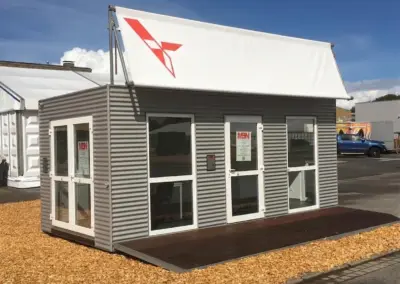 Silbergrauer Eventcontainer mit Wellblechverkleidung, weißen Fenstern und Türen, dunkler Holzterrasse und einem weißen Dachbanner. Der Showroom-Container steht auf einem mit Holzhackschnitzeln ausgelegten Messestand bei sonnigem Wetter.