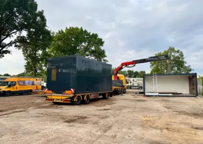 Eine detaillierte Außenaufnahme zeigt den Aufbau einer modernen, anthrazit farbenen MBN-Containeranlage. Ein gelber MBN-Kran-LKW mit ausgefahrenem rotem Kranarm hebt ein Bürocontainer-Modul präzise auf ein Fundamentmodul. Im Hintergrund stehen Bäume, ein LKW-Anhänger mit einem weiteren Modul und ein gelber MBN-Lieferwagen.