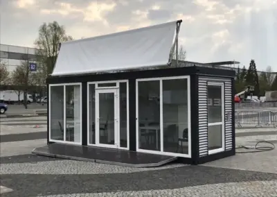 Moderner Event- und Showroom-Container von MBN mit schwarzem Stahlrahmen, großflächigen Glasfenstern, weißem Dachbanner und integrierter Holzterrasse auf einem gepflasterten Veranstaltungsgelände.