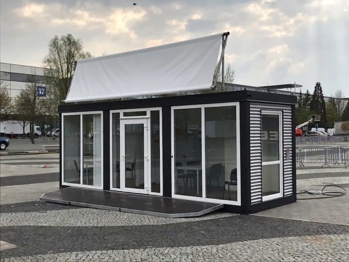 Moderner Event- und Showroom-Container von MBN mit schwarzem Stahlrahmen, großflächigen Glasfenstern, weißem Dachbanner und integrierter Holzterrasse auf einem gepflasterten Veranstaltungsgelände.