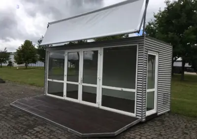 Freistehender Eventcontainer mit silbergrauer Wellblechfassade, weiß gerahmten Glasfenstern, dunkler Holzterrasse und einem Dachbanner auf einem gepflasterten Außenbereich.