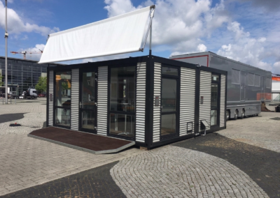 moderne Containeranlage von MBN in Wellblech-Optik mit schwarzem Rahmen, bodentiefen Glastüren und blankem Werbebanner auf einem Messegelände.