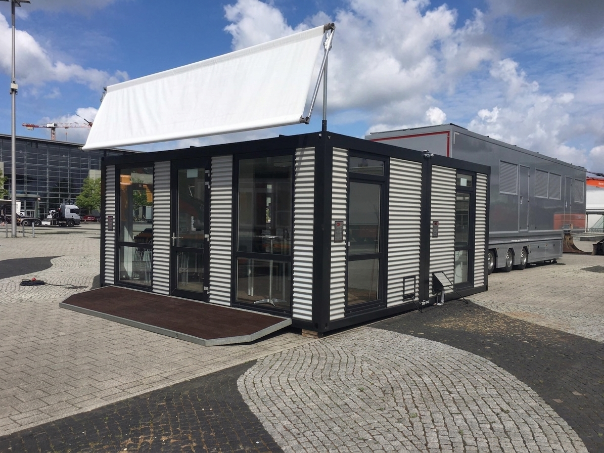 moderne Containeranlage von MBN in Wellblech-Optik mit schwarzem Rahmen, bodentiefen Glastüren und blankem Werbebanner auf einem Messegelände.