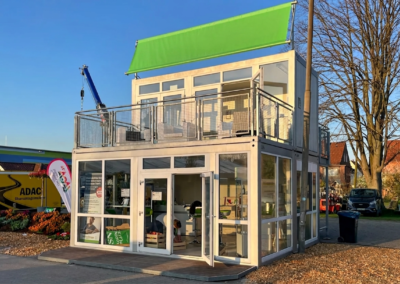 Zweistöckiger modularer Containerbau von MBN mit vollflächiger Glasfassade im Erdgeschoss, Balkon mit Metallgeländer und einem blanken, grünen Werbebanner oben.